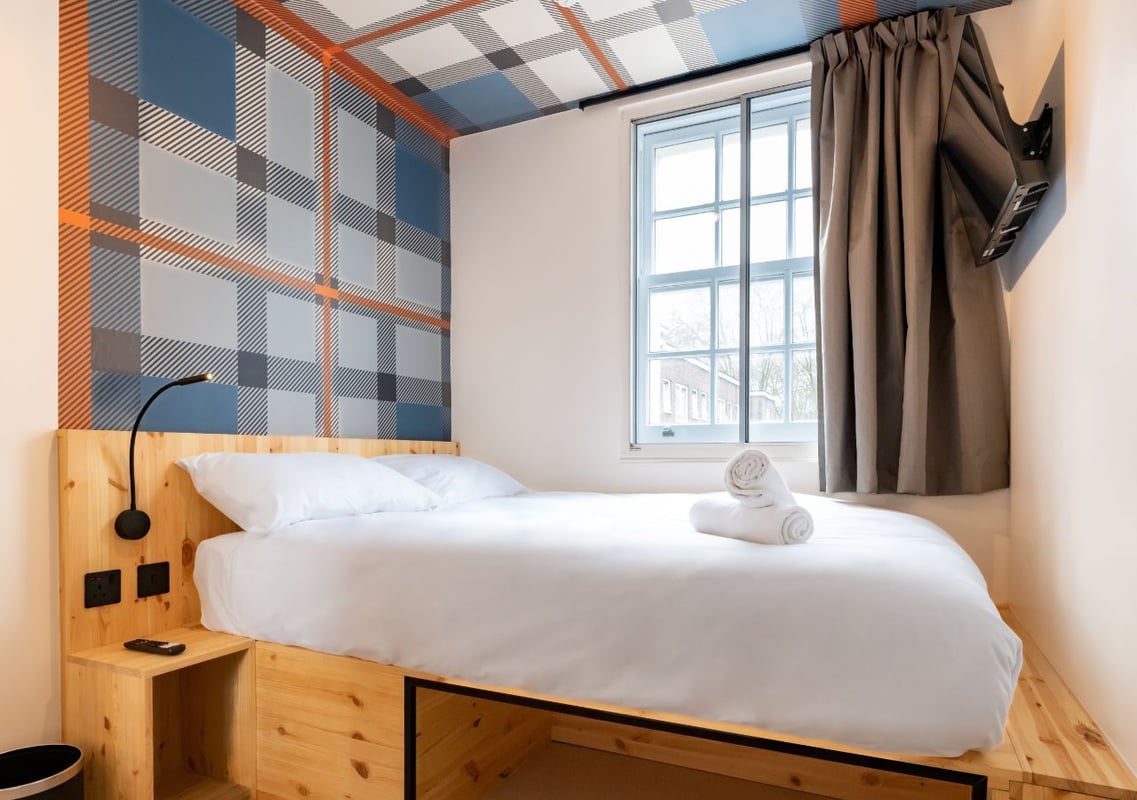 Compact easyHotel Paddington double room featuring a wooden platform bed with white bedding, a distinctive blue, grey, and orange plaid accent wall and ceiling, and a window with curtains.
