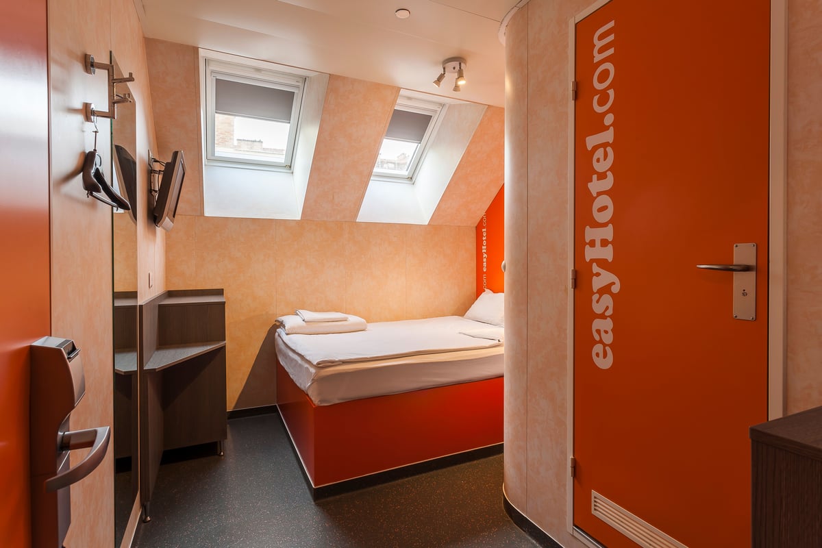 Compact easyHotel single room with a modern orange bed, white bedding, and two skylight windows under a sloped ceiling. The room features minimalist decor, a wall-mounted TV, and easyHotel branding on the door.