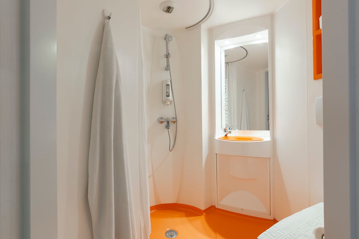 A minimalist easyHotel room bathroom featuring a bright orange wet room shower floor, a compact sink with an orange basin, a mirror, and a white towel hanging on a hook. The efficient design is typical of a standard easyHotel room.