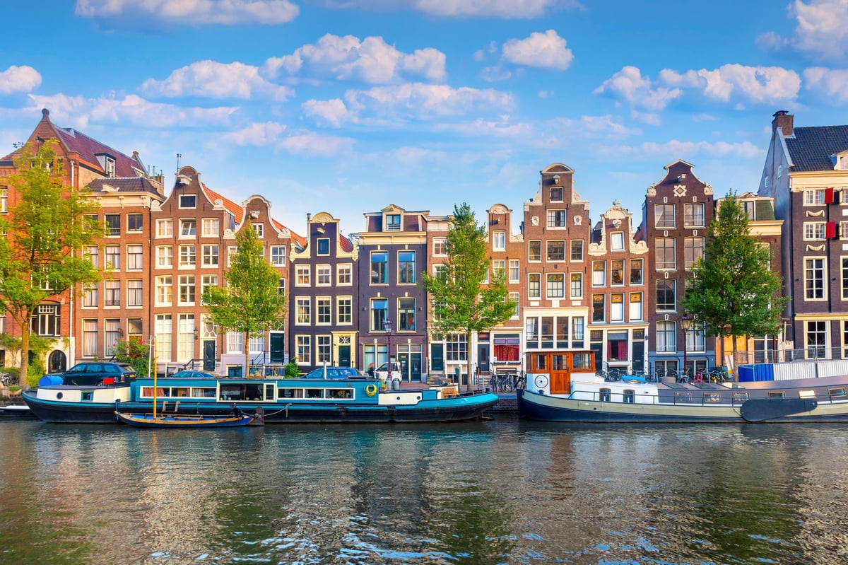 A vibrant canal scene in Amsterdam city centre featuring historic gabled houses, green trees, and multiple houseboats docked along the water under a clear blue sky with white clouds.