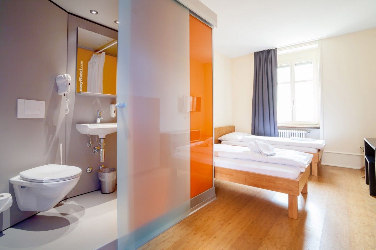 Modern easyHotel room in Basel with two single beds, wooden flooring, and a compact en-suite bathroom featuring a frosted glass and orange sliding door.