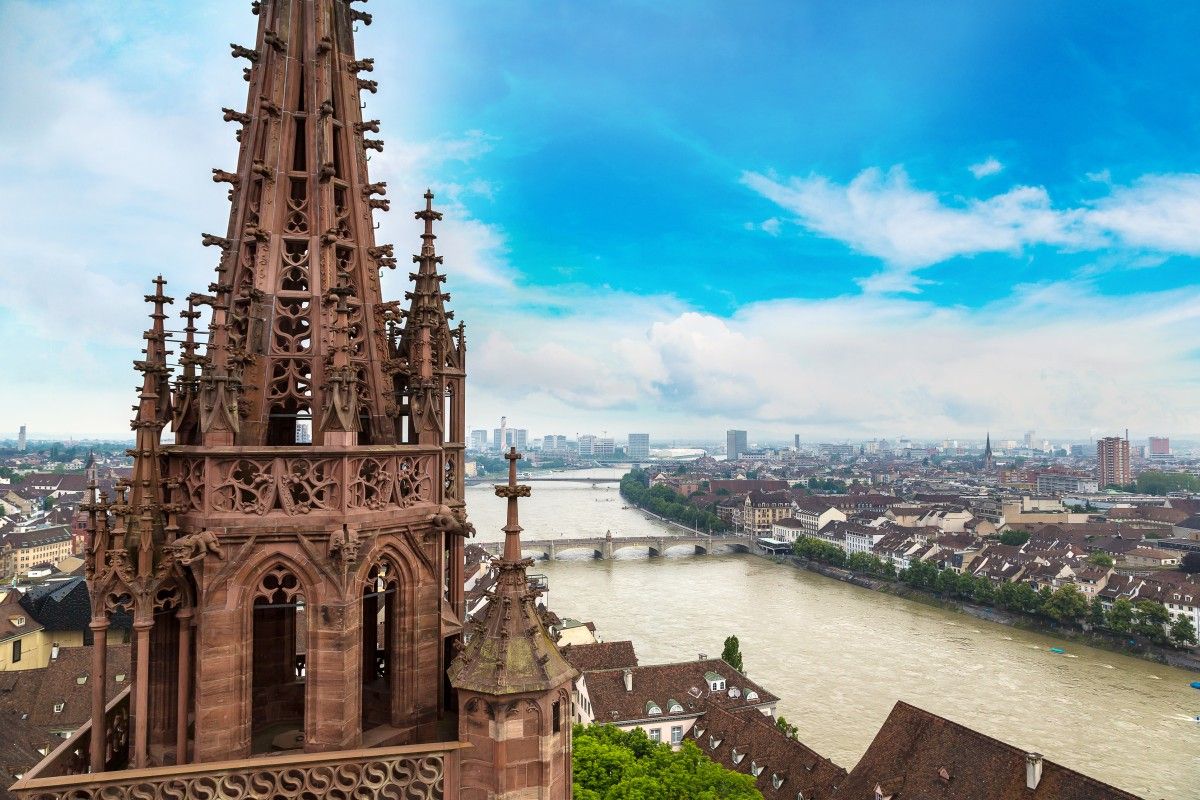 Panoramic view of Basel, Switzerland, from the Basel Minster, featuring its intricate red sandstone Gothic spire, the Rhine River winding through the city, and a distant urban skyline under a blue sky.