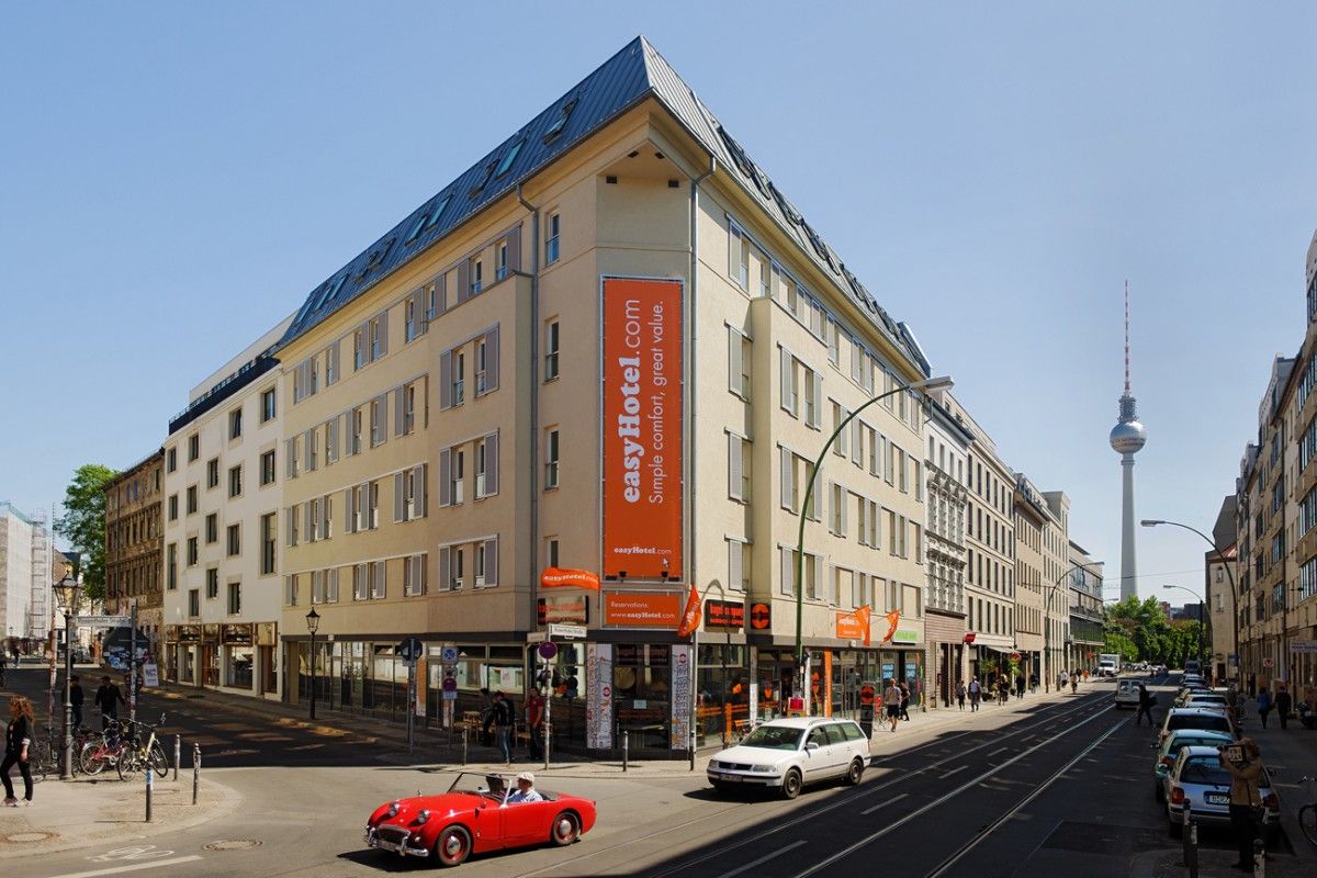 Exterior view of the easyHotel Berlin building on a sunny day, located on a city street with pedestrians, cars, and the iconic Berliner Fernsehturm visible in the background.