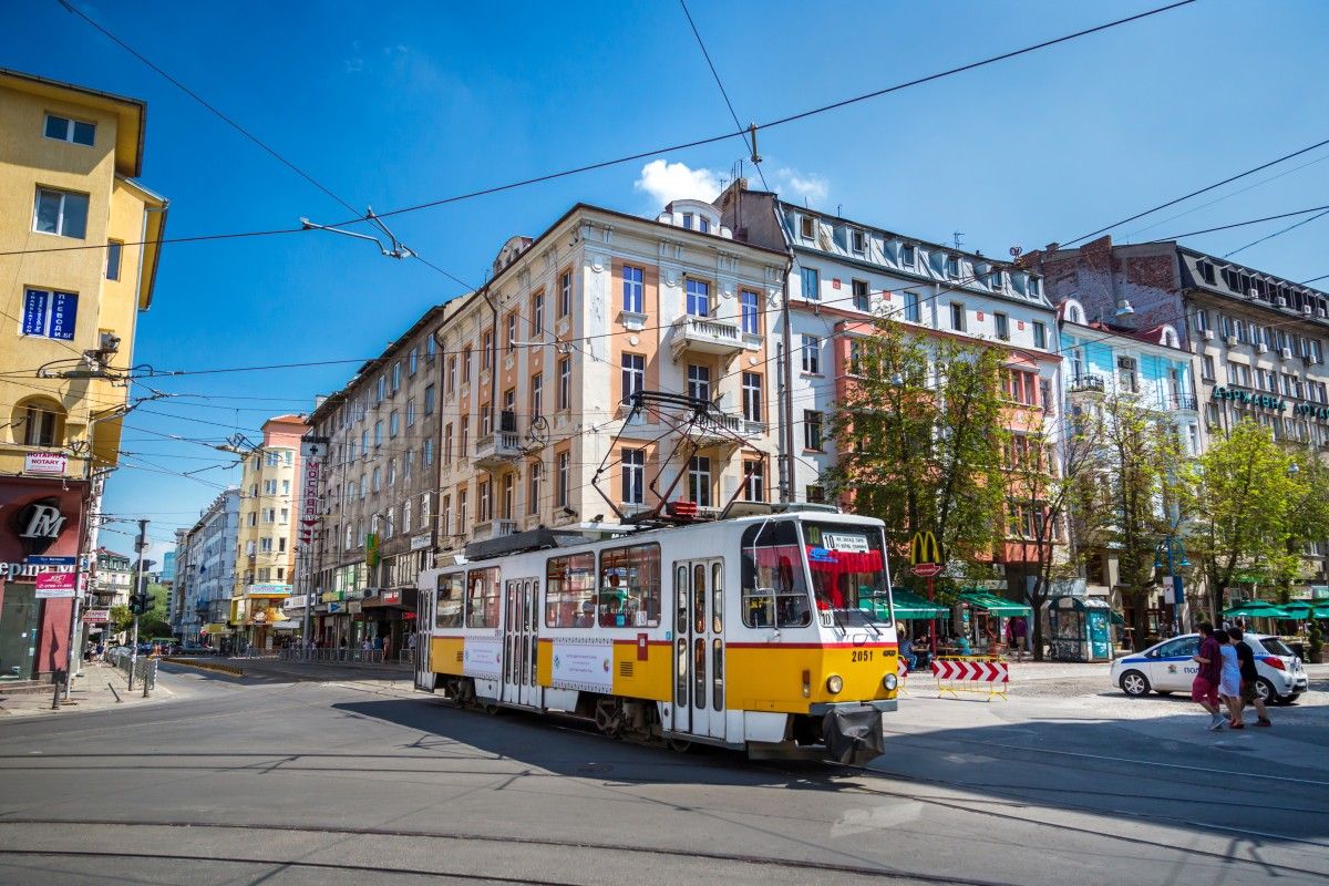 Yellow and white tram traveling on a street in Sofia, Bulgaria, with historic city buildings and a clear blue sky.