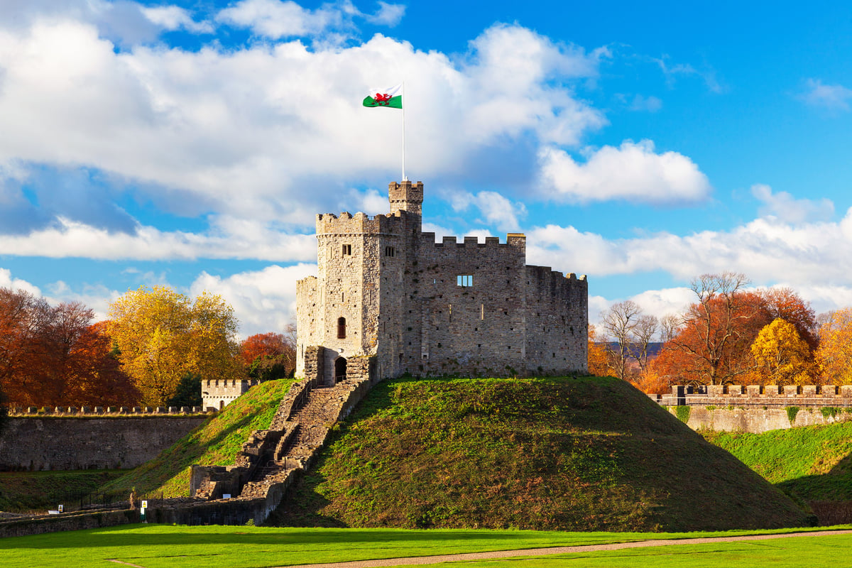 Cardiff Castle's Norman Keep on a grassy motte, with the Welsh flag flying under a bright blue sky and vibrant autumn trees surrounding the historic landmark.
