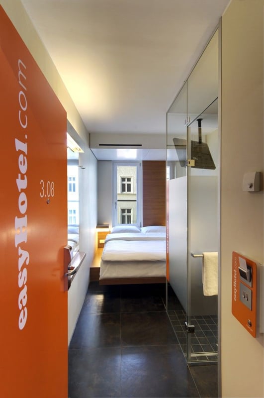 Compact easyHotel room in Berlin with a bright orange door, double bed with white linens, dark tiled floor, and a modern glass shower enclosure.