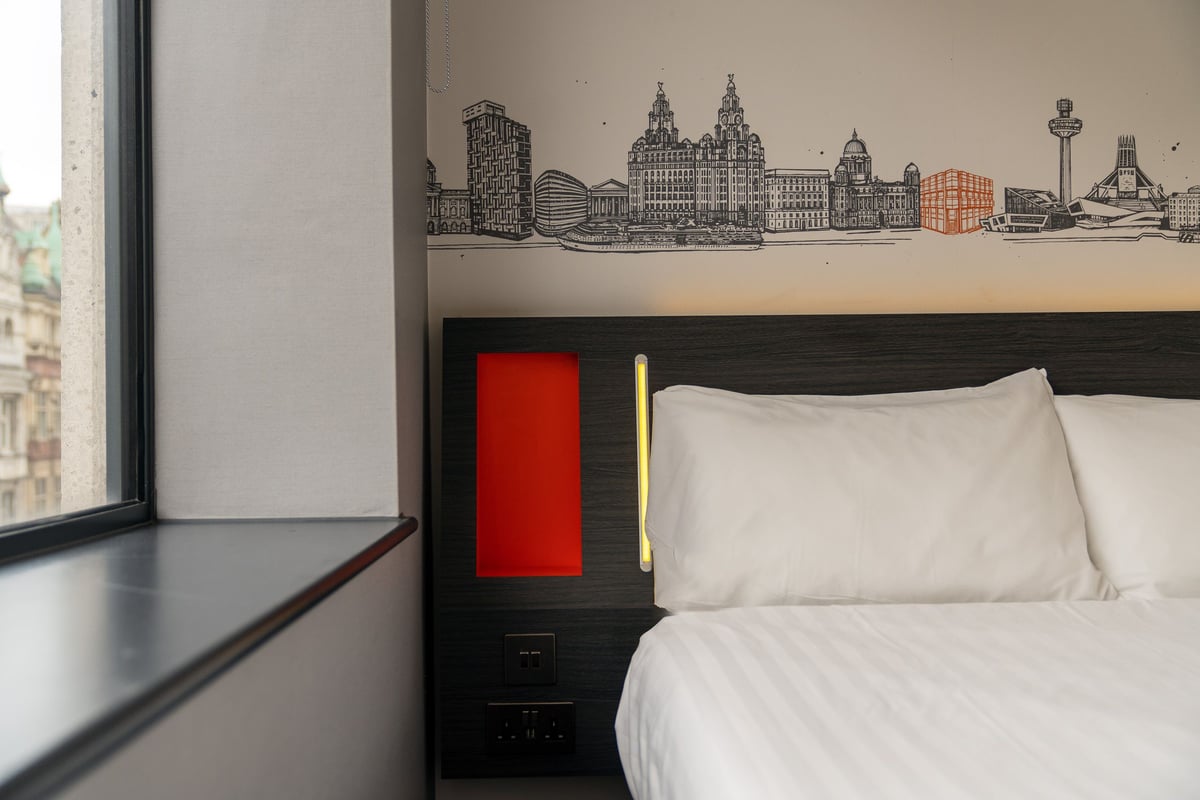 A modern easyHotel double room in Liverpool, featuring a distinctive black and white mural of the city's iconic skyline above the bed. The room has crisp white bedding, a dark headboard with integrated red and yellow lighting, and a large window offering 