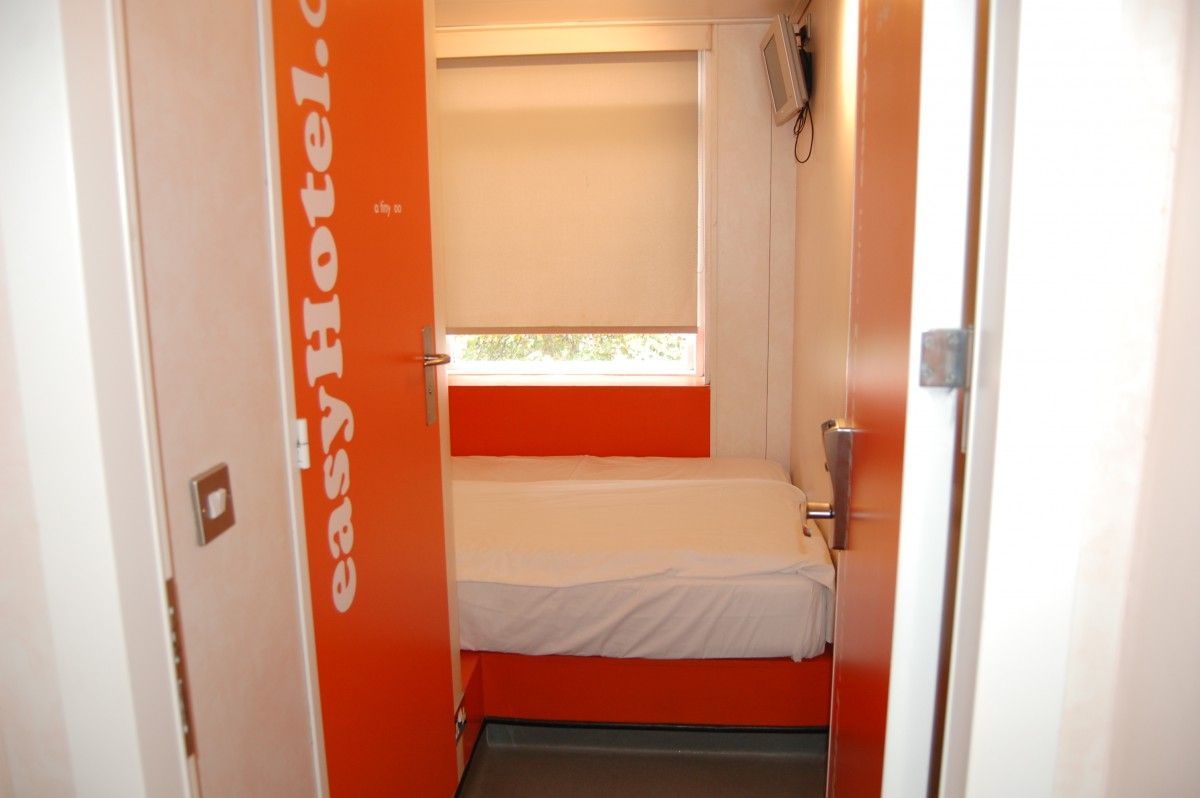 A compact easyHotel room featuring a double bed on an orange platform, a window, and a wall-mounted TV, viewed through the partially open signature orange easyHotel branded door.