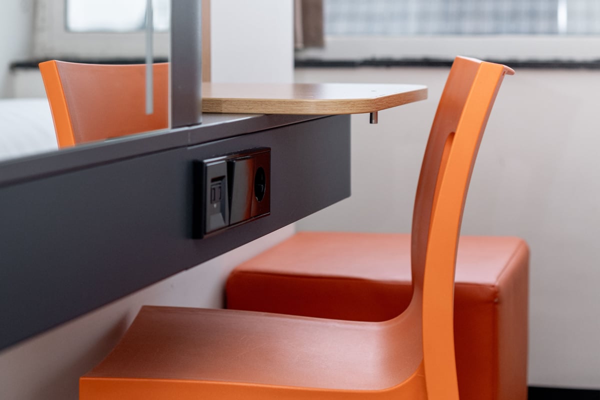A modern desk area in an easyHotel Amsterdam Arena Superior Double room, featuring a vibrant orange chair and ottoman, a dark grey vanity with a wooden shelf, and convenient built-in power and USB outlets.