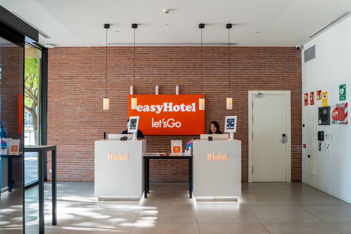 easyHotel Barcelona Fira reception desk with staff, brick wall, and orange signage.