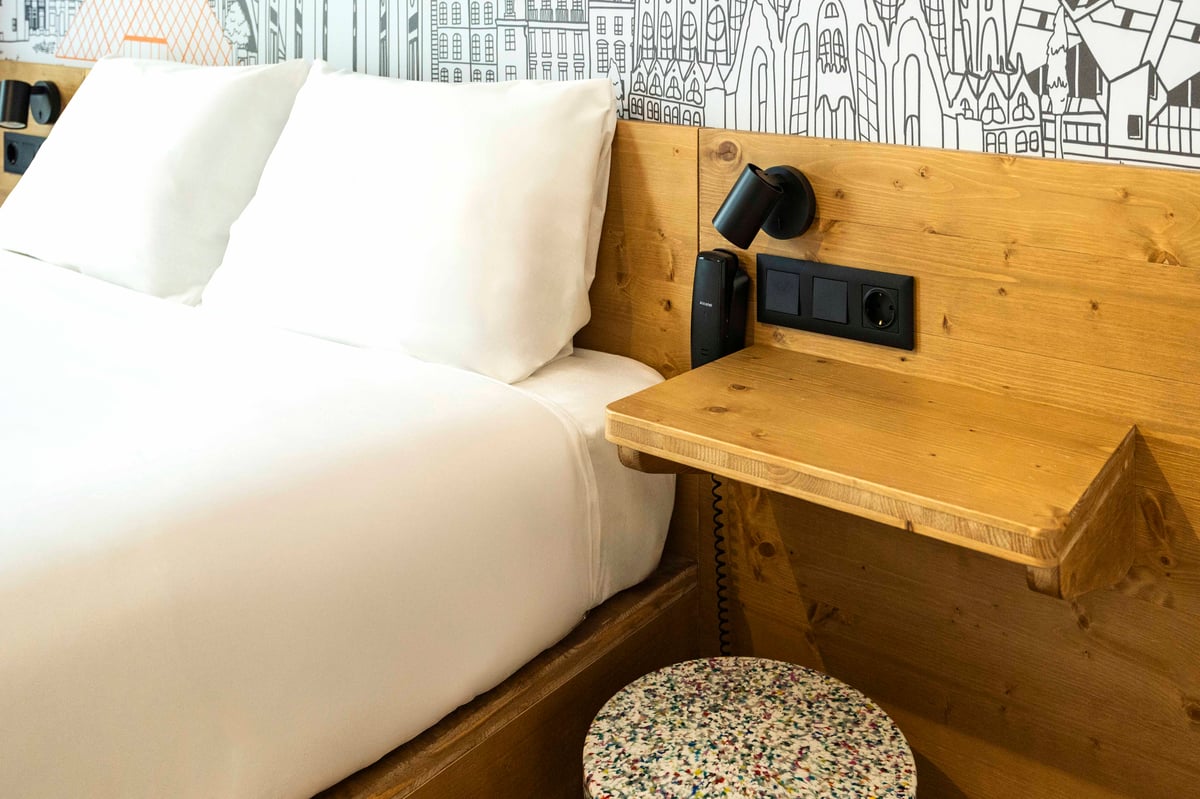 EasyHotel Barcelona La Sagrera double room with white linens, pillows, and a wooden headboard featuring a built-in desk and wall-mounted reading lamp.