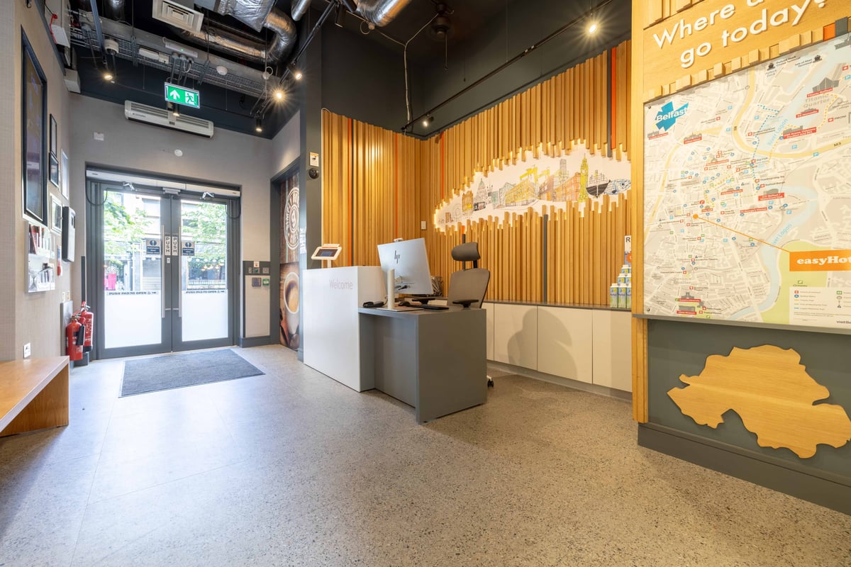 Modern easyHotel Belfast reception area with a white front desk and computer, featuring a large wooden slatted wall with a colorful city skyline mural, and a prominent Belfast map.