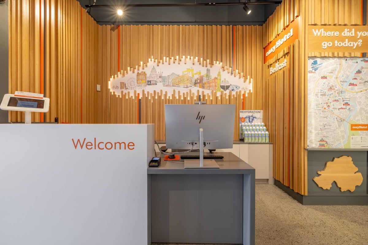 easyHotel Belfast reception desk with a welcome sign, modern wood-paneled wall featuring a Belfast cityscape mural, and an elevator.