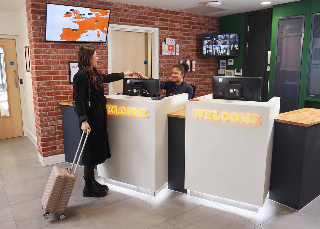 Reception desk with a guest checking in, featuring a large screen, computer, and welcome signage in a modern, brick-accented lobby.