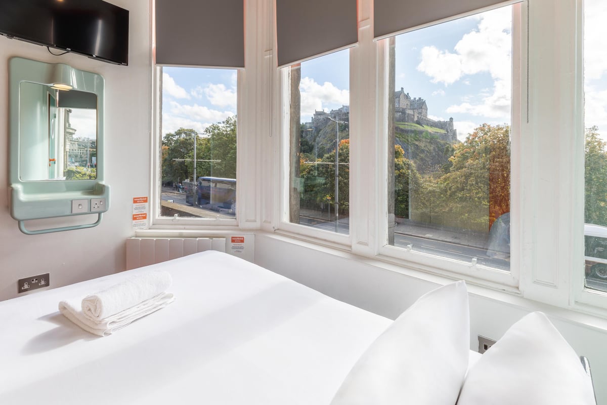 easyHotel Edinburgh double room with a crisp white bed, a signature light blue mirror, and a panoramic window view of Edinburgh Castle on a sunny day.
