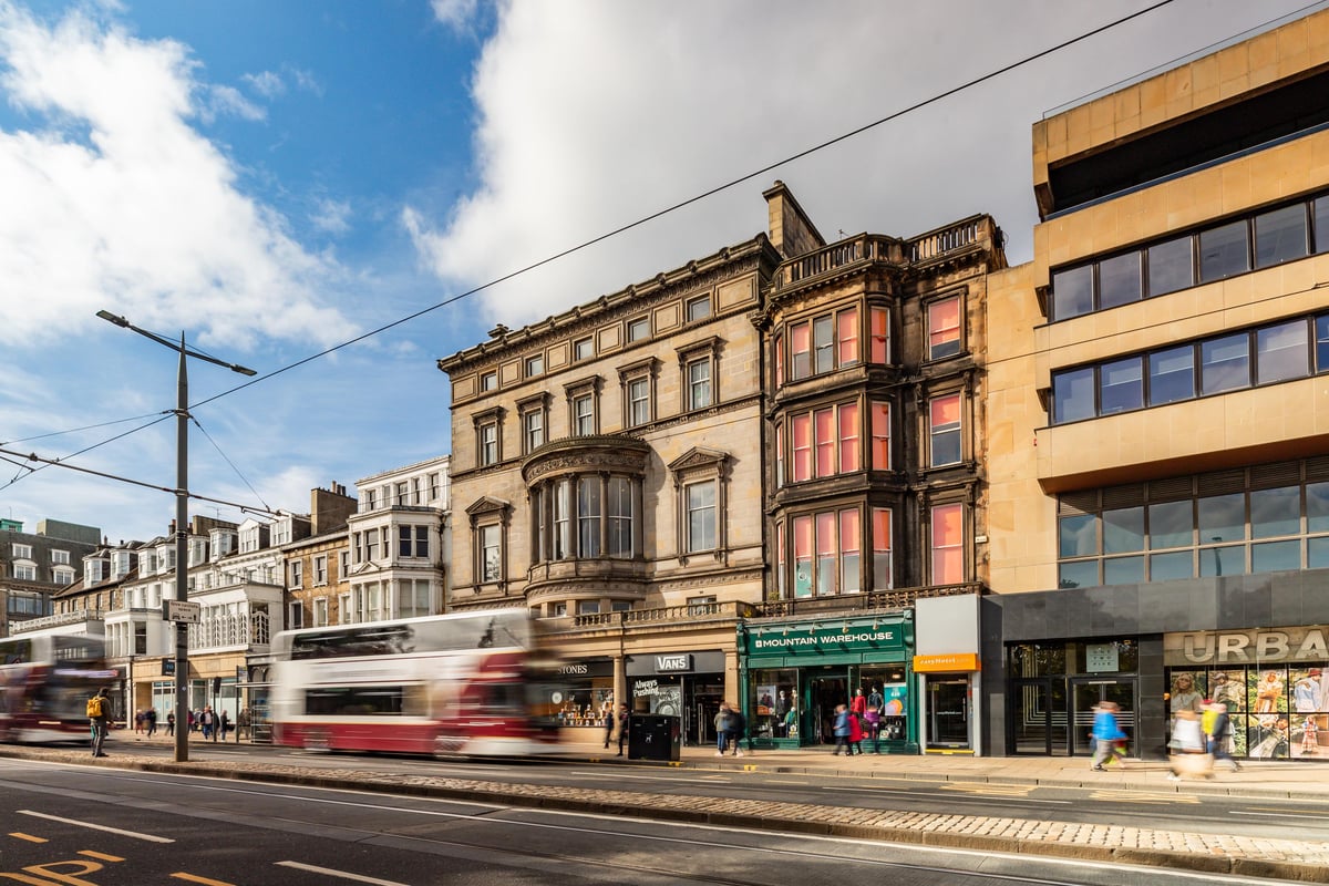 A vibrant street scene in Edinburgh, featuring classic and modern buildings, including the easyHotel Edinburgh exterior. Red double-decker buses pass by pedestrians and shops like Mountain Warehouse under a clear blue sky.
