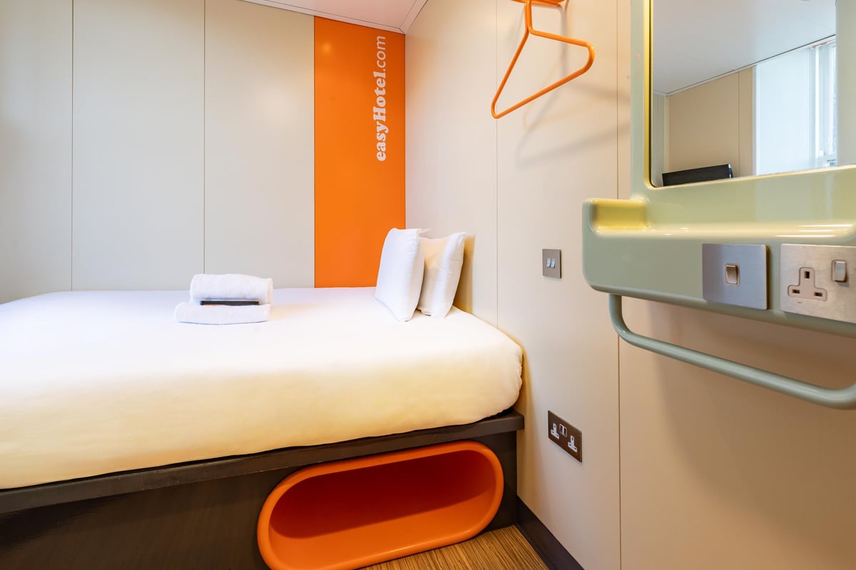 A compact small double room at easyHotel Edinburgh, featuring a bed with white linen, an orange easyHotel branded accent wall, and a light green integrated sink with mirror and power outlets.