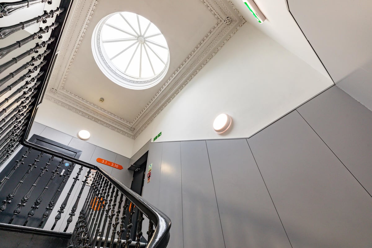 A brightly lit easyHotel Edinburgh staircase featuring an ornate black metal railing, a prominent circular skylight, and contemporary grey wall panelling leading to rooms 3.1-3.10.