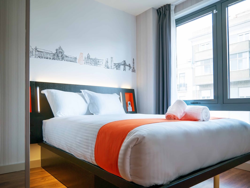 Modern easyHotel Lisbon double room featuring a crisp white bed with an orange runner, a decorative Lisbon city skyline mural on the wall, and ample natural light from a large window.