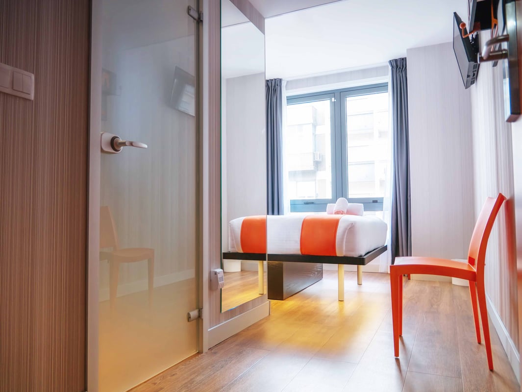 Modern easyHotel Lisbon double room with a white bed featuring orange accents, an orange chair, hardwood floors, large windows with gray curtains, and a wall-mounted TV.