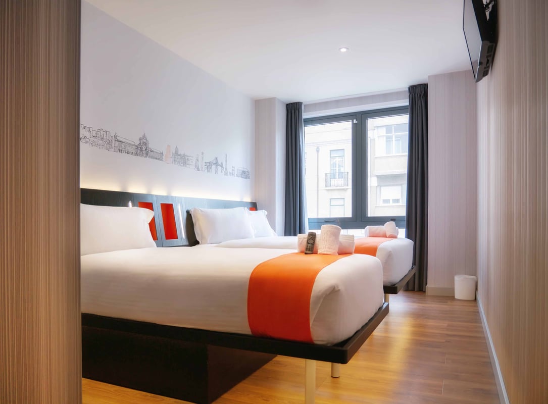 easyHotel Lisbon family triple room with three beds, white bedding, orange accents, and a city skyline mural above the headboard.