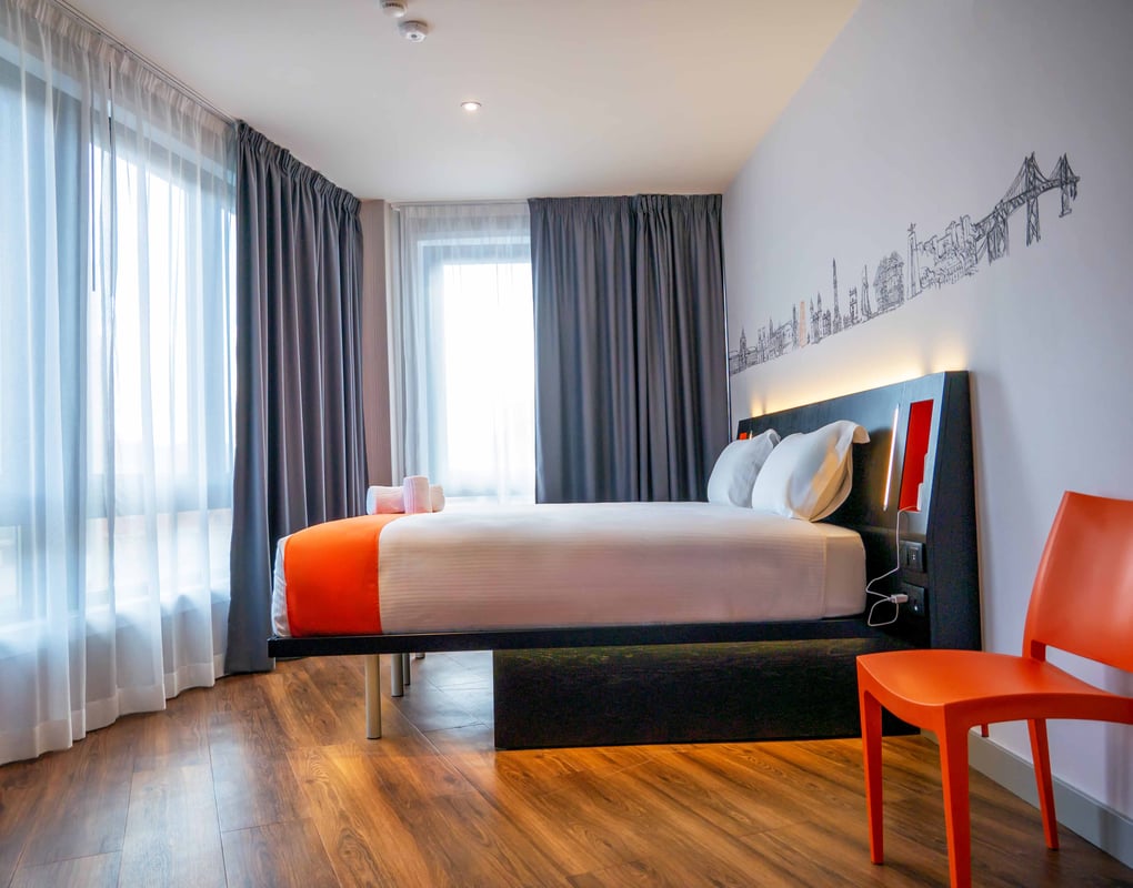 An easyHotel Lisbon Superior Double room featuring a comfortable double bed with white linen and an orange accent, a bright orange chair, and a Lisbon city skyline mural above the bed. The room has light wooden flooring and large windows with sheer and bl