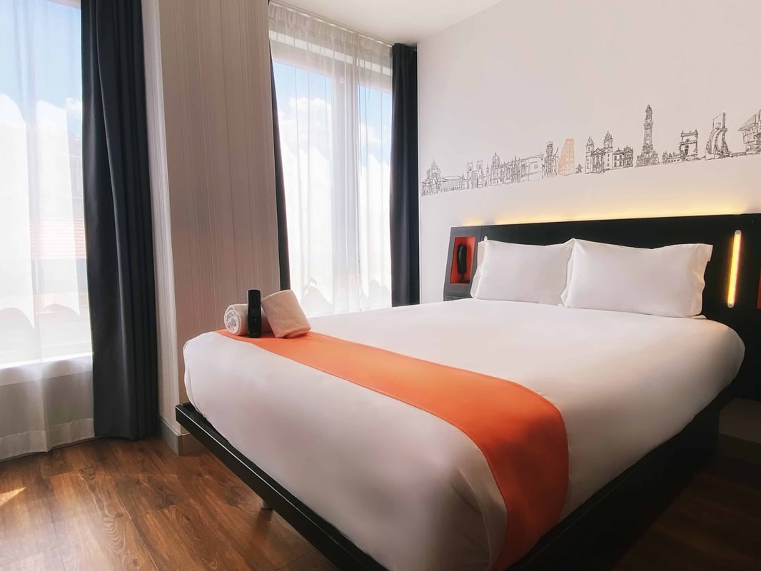 easyHotel Lisbon superior double room with a modern design, featuring a large bed with white linen and orange runner, a dark headboard with integrated lighting, and a wall mural depicting iconic Lisbon landmarks. The room has wood-effect flooring and larg
