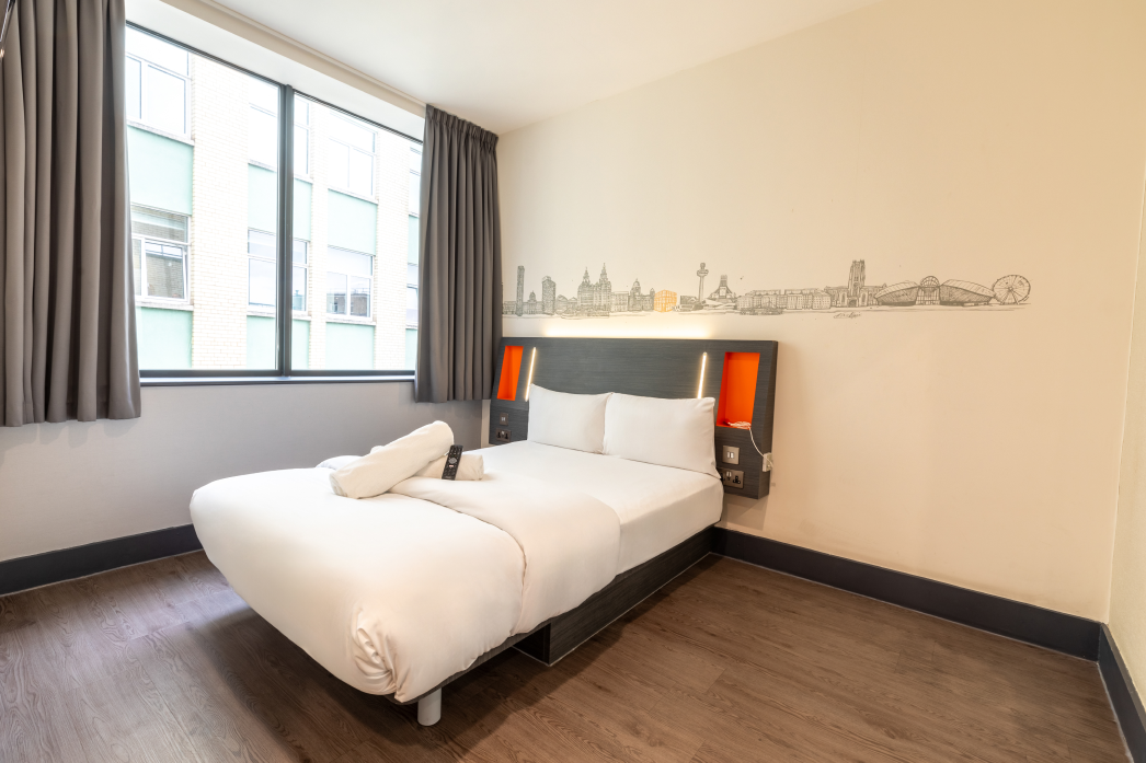Spacious easyHotel Liverpool accessible room featuring a comfortable double bed, a large window with natural light, and a stylish wall mural depicting the iconic Liverpool skyline above the bed's headboard.