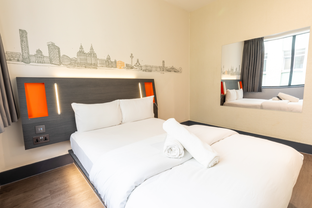 An easyHotel accessible room in Liverpool featuring a comfortable bed with white bedding, a modern headboard with orange details and integrated lighting, and a distinct mural of the Liverpool city skyline on the wall.