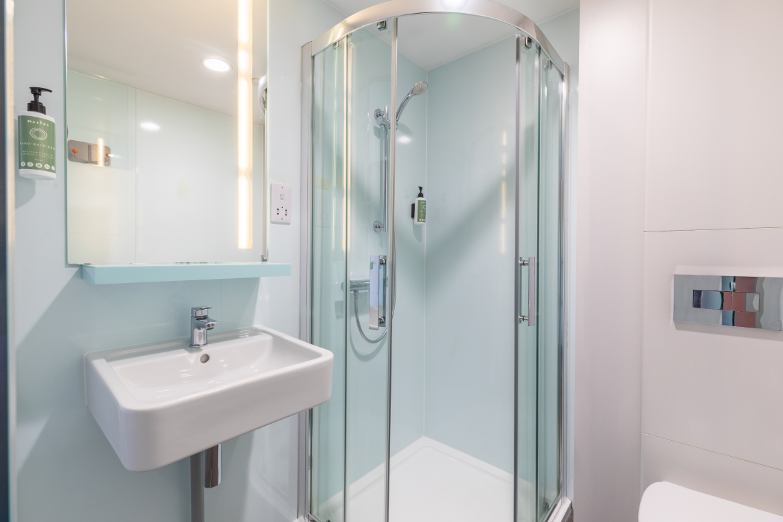 A compact, modern easyHotel Liverpool bathroom featuring a white sink, large mirror with integrated light, and a clear glass shower enclosure.