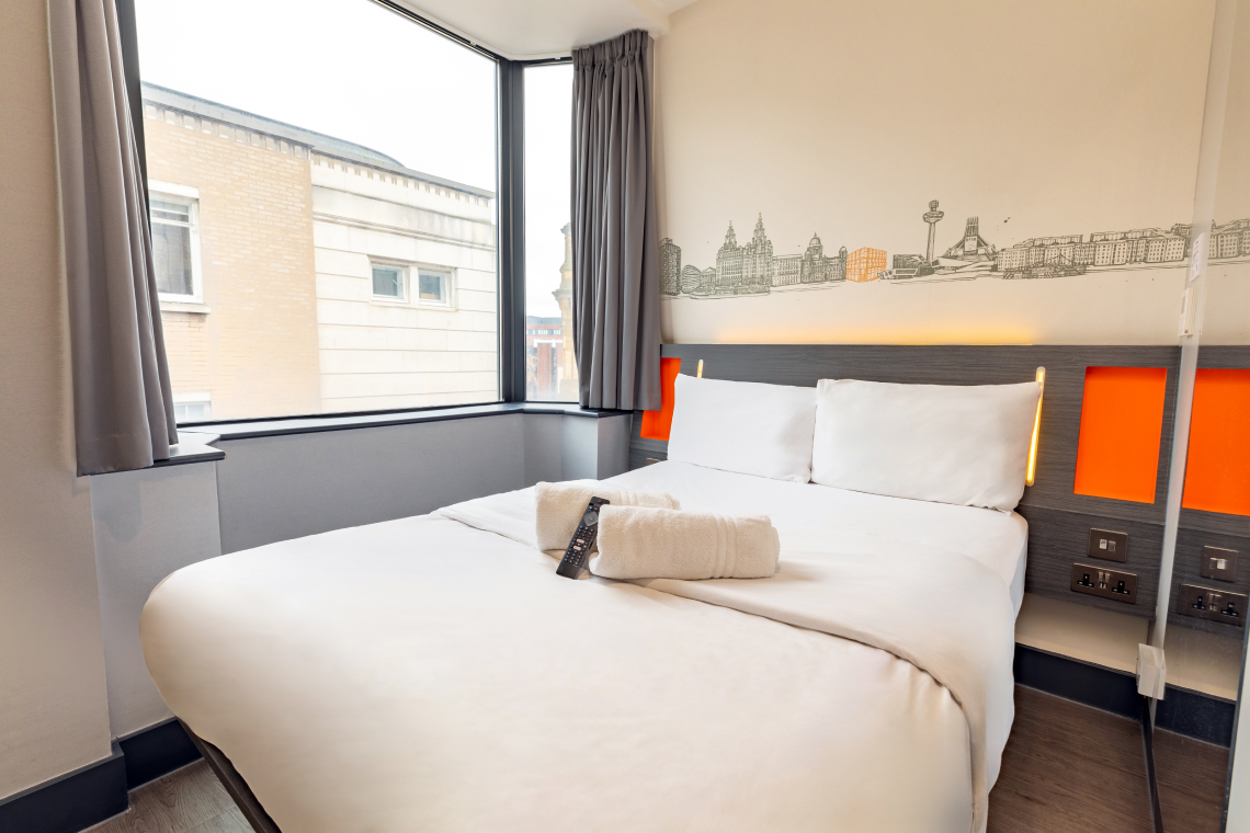 Modern easyHotel Liverpool double room with a clean white bed, rolled towels, and a distinctive Liverpool skyline mural above the headboard, next to a corner window.