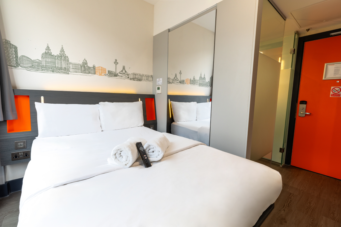 EasyHotel Liverpool double room featuring a clean, white double bed, vibrant orange accents, and a distinctive Liverpool skyline mural above the headboard.