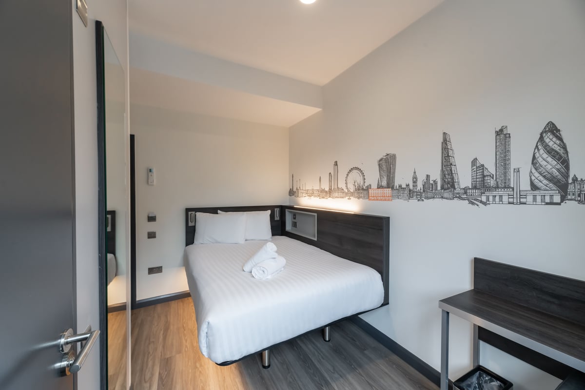 easyHotel London Victoria Standard Double Room featuring a white double bed, dark wood furniture, and a distinctive London skyline wall mural.