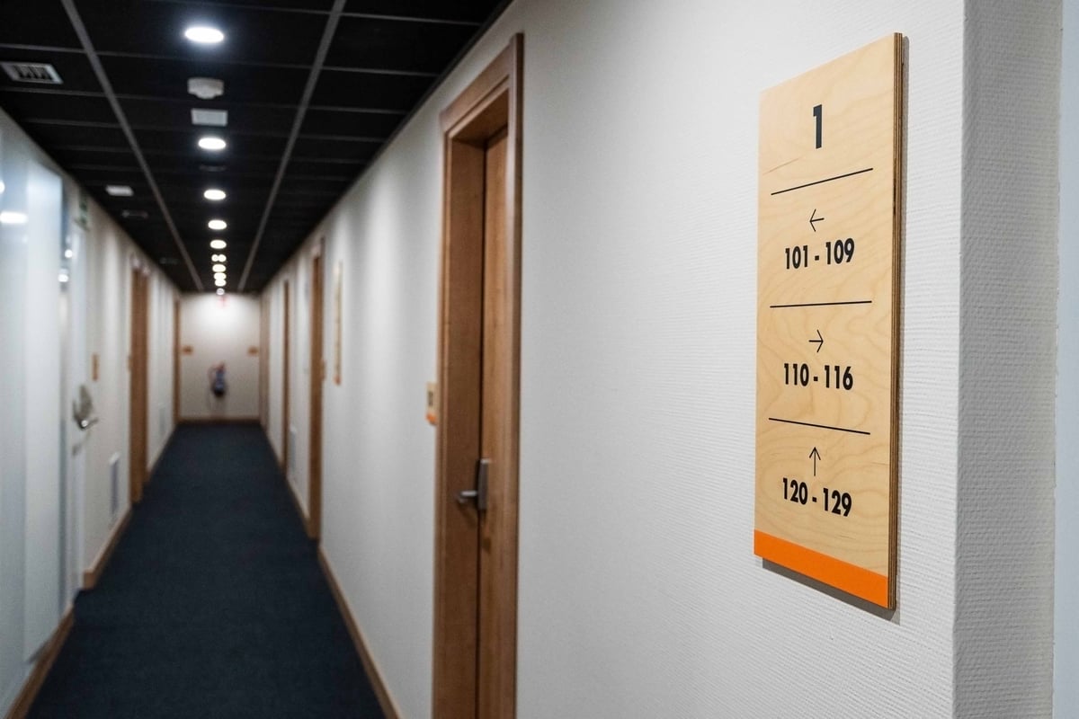 First-floor corridor at easyHotel Madrid Alcalá, featuring a long, bright hallway with wooden room doors, a dark blue carpet, and a prominent wooden sign displaying room numbers 101-129 with directional arrows.