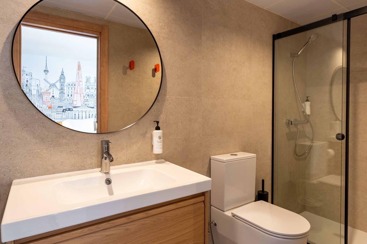 A modern easyHotel Madrid Alcalá bathroom with beige tiled walls. It features a white sink on a light wooden vanity, a large round mirror reflecting a graphic city skyline mural, a white toilet, and a walk-in shower with a glass door and black frame.
