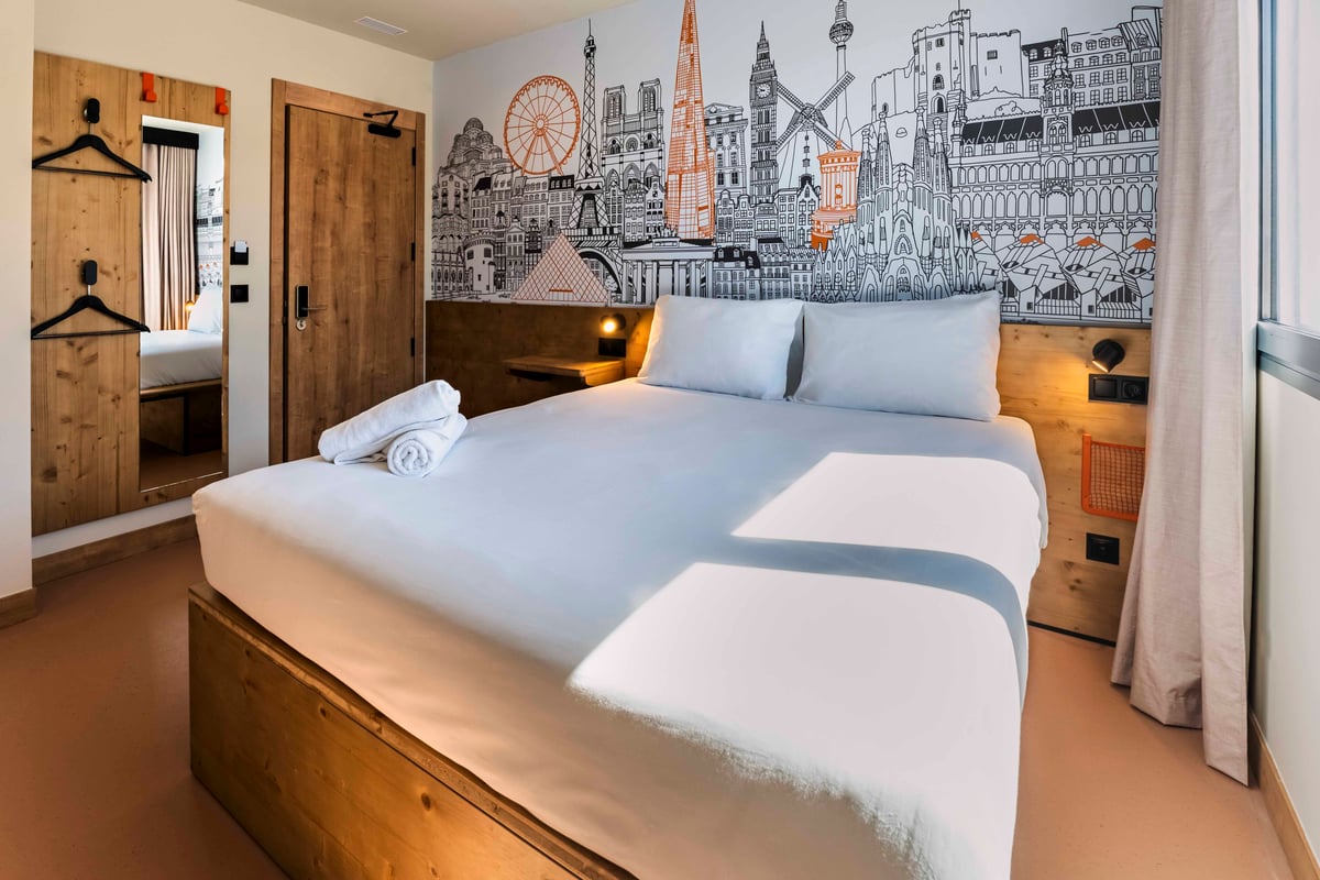 An easyHotel Madrid Alcalá double bedroom with a white-linened bed and a prominent mural of European city landmarks on the wall. The room features practical wooden furnishings, including a headboard with a small shelf and integrated lighting, a wooden pan