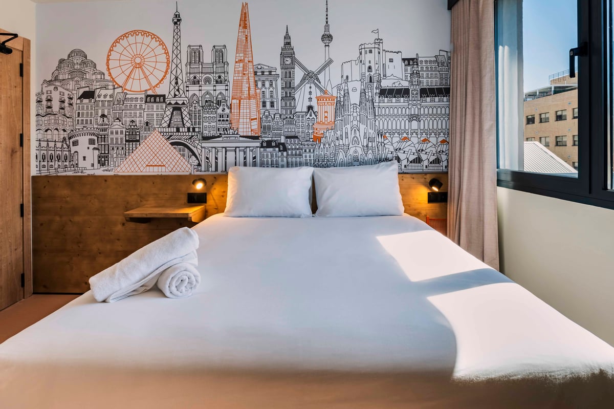 easyHotel Madrid Alcalá double bedroom featuring a white bed, wooden headboard with built-in lights, and a vibrant mural depicting famous European city landmarks. Rolled white towels are placed on the bed, with natural light illuminating the room from the