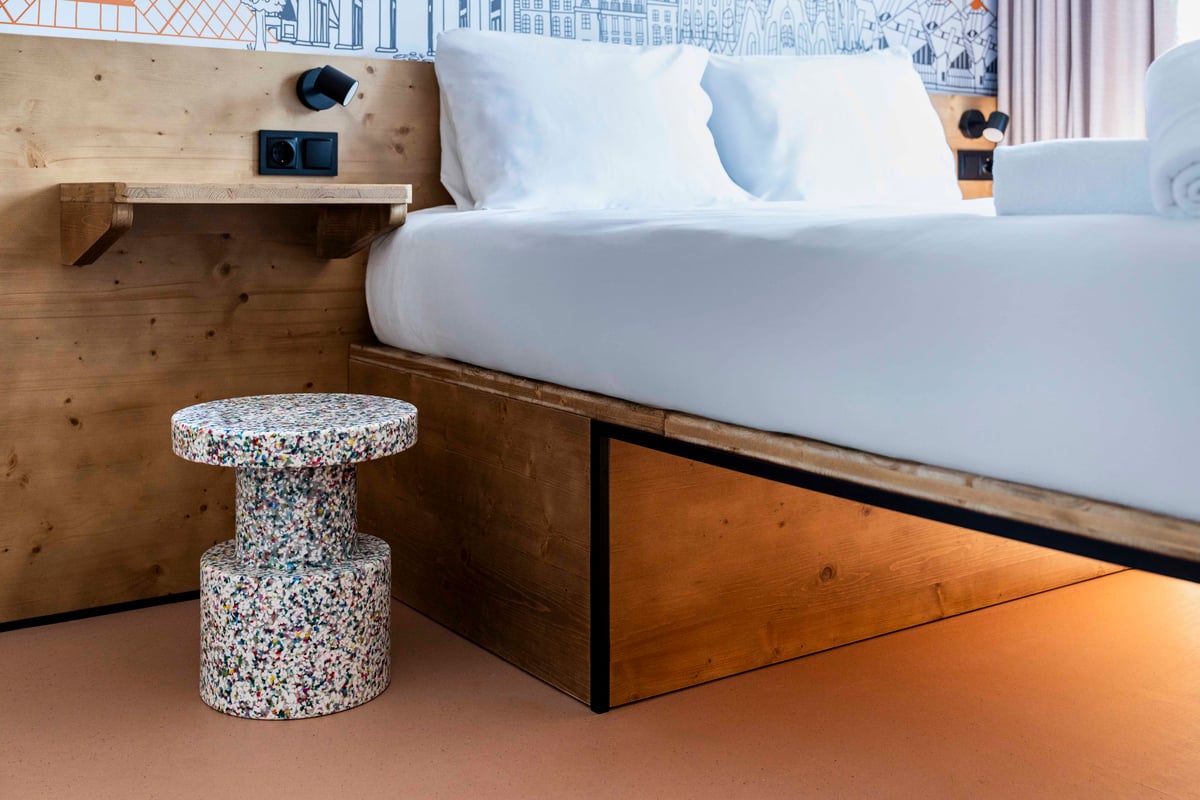 easyHotel Madrid Alcalá double bedroom featuring a crisp white bed, wooden headboard with integrated shelving and bedside lamp, and a unique multicolored speckled stool. Warm under-bed lighting highlights the modern, functional design.