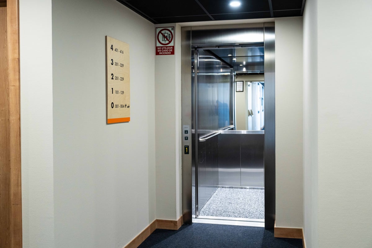 A modern, stainless steel elevator with open doors in a well-lit hallway at easyHotel Madrid Alcalá. A wooden sign on the wall indicates floor numbers and room ranges (0-4), accompanied by a red "no utilizar en caso de incendio" (do not use in case of fir
