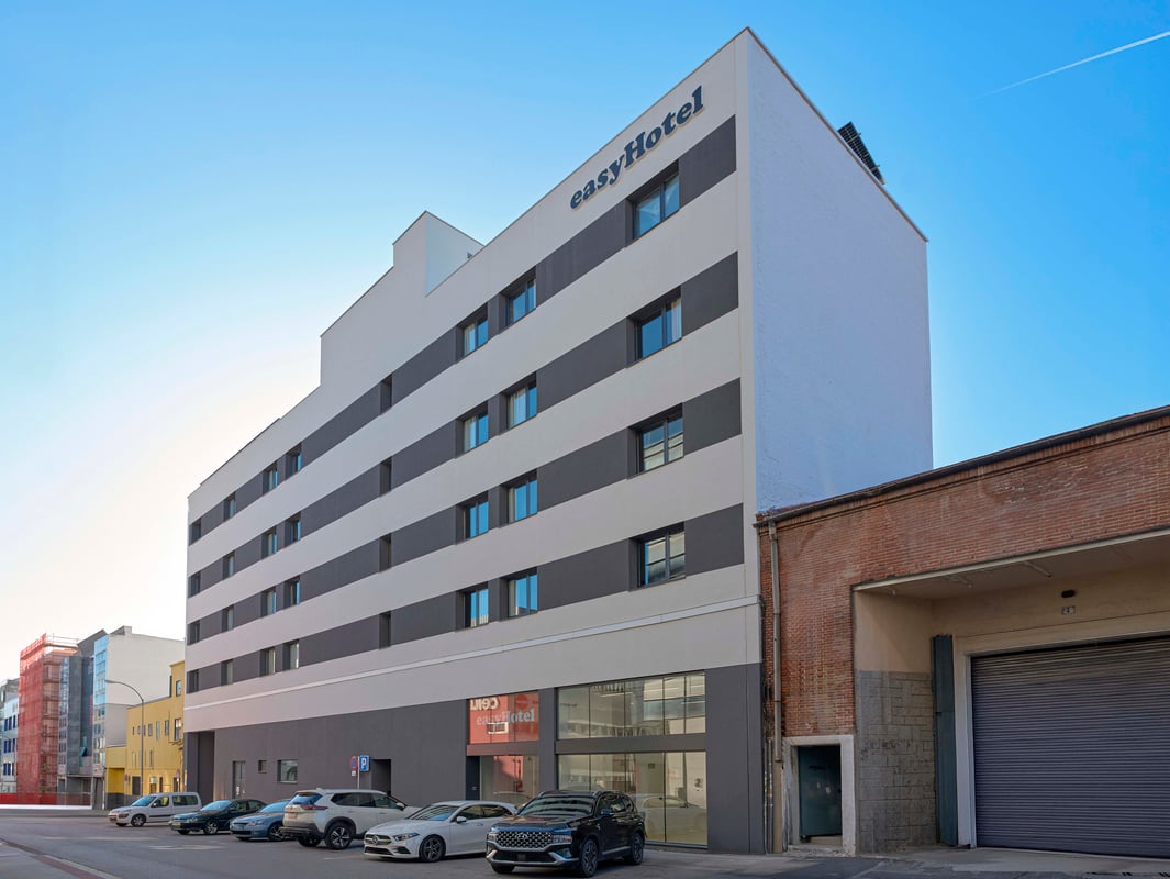 Exterior view of the easyHotel Madrid Alcala, a modern building with a distinctive white and dark grey striped facade, cars parked in front, and a clear blue sky.