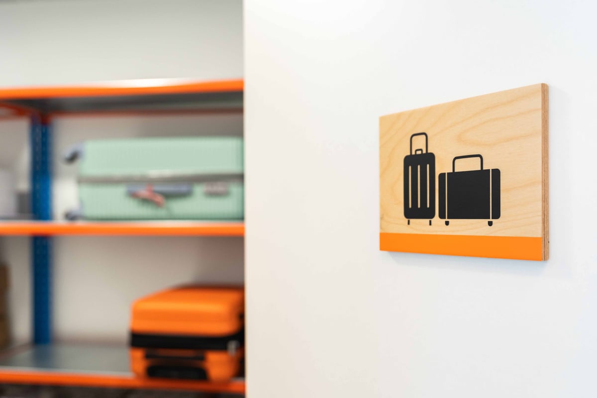 Luggage storage sign at easyHotel Madrid Alcalá featuring black suitcase icons on a wooden plaque with an orange stripe, positioned above blurred shelves holding various suitcases.