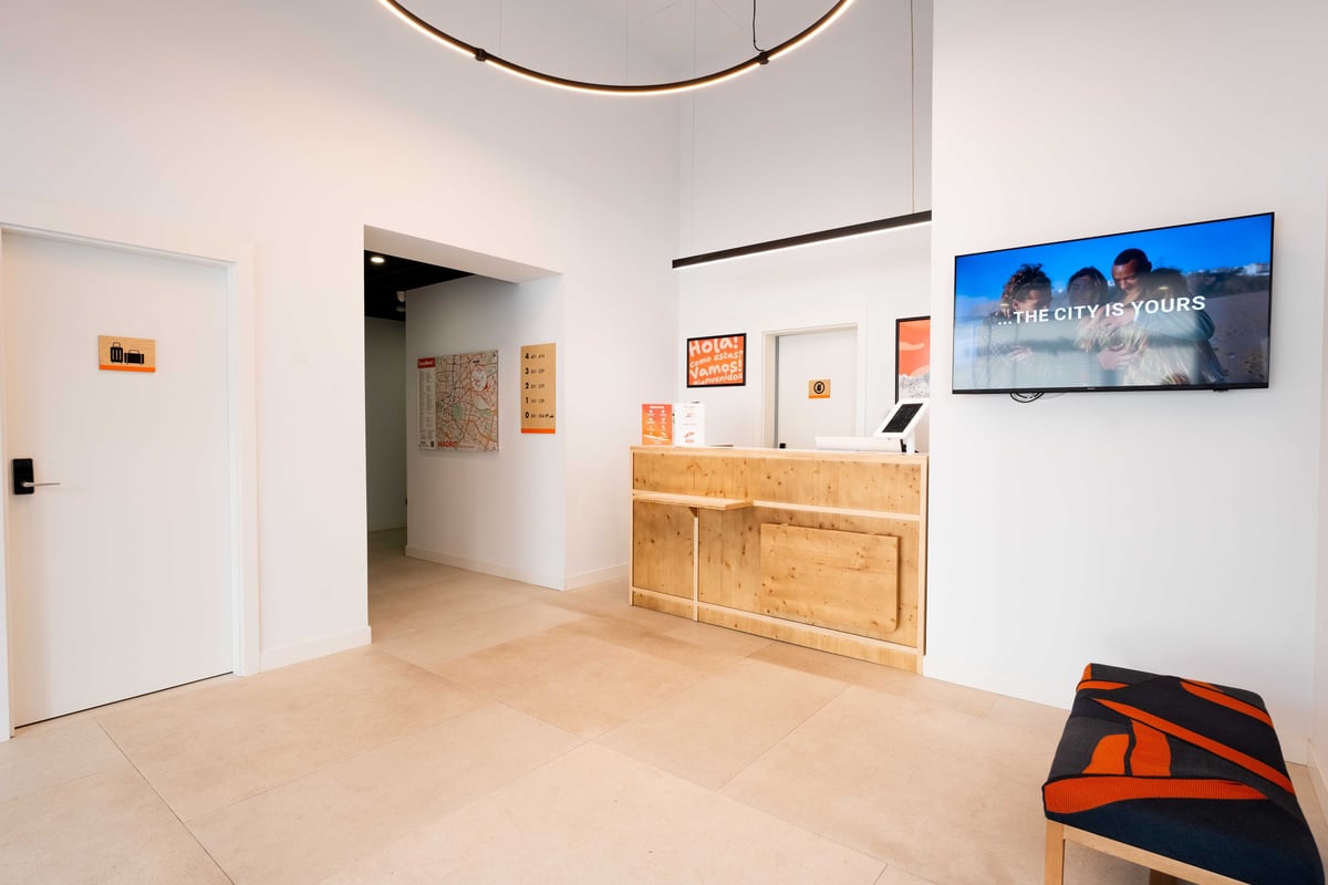 easyHotel Madrid Alcalá reception area with a wooden reception desk, a mounted television displaying an advertisement, and a seating area.