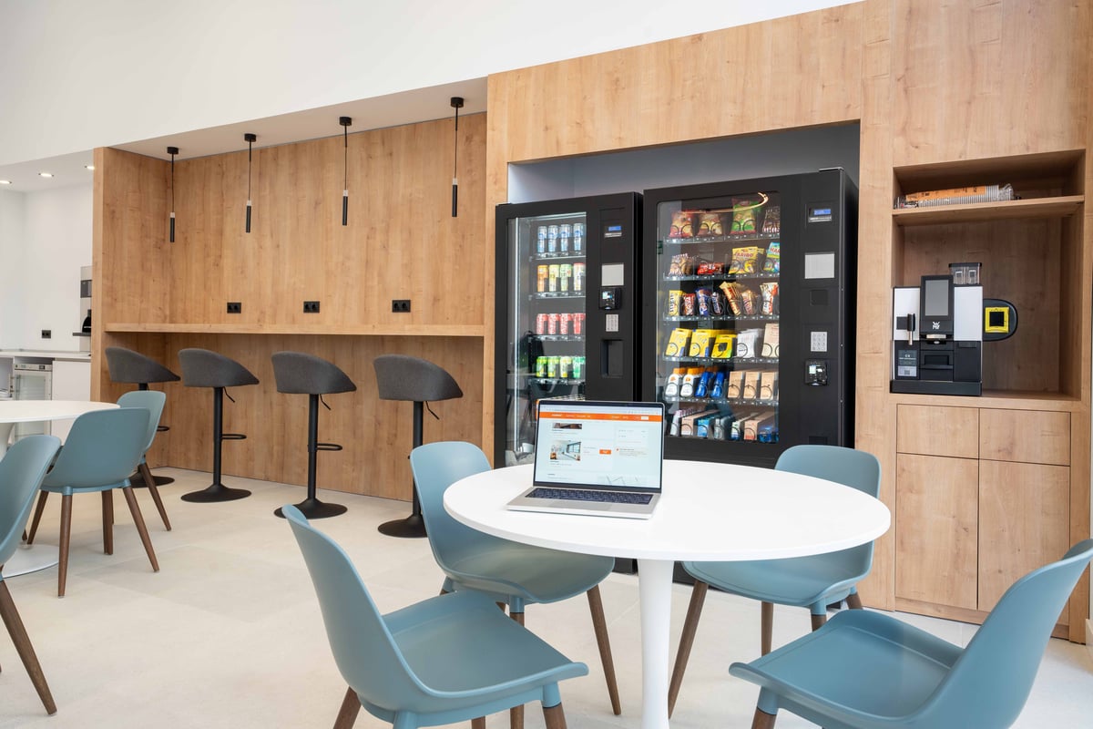 easyHotel Madrid Alcalá self-service refreshment area with two vending machines for drinks and snacks, a coffee machine, a long wooden counter with bar stools, and round white tables with light blue chairs, designed for guest convenience.