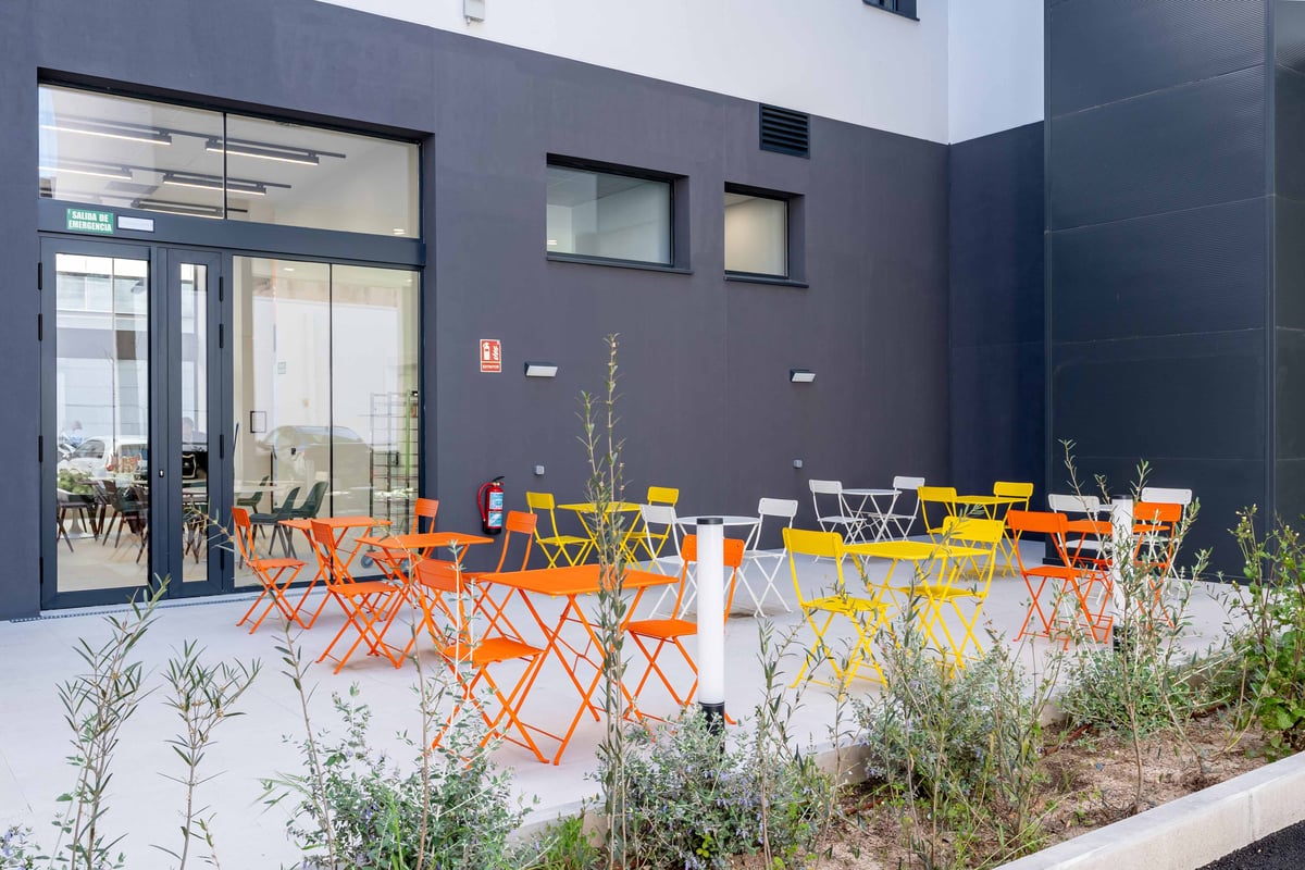 easyHotel Madrid Alcalá terrace with colorful orange, yellow, and white outdoor seating. The modern patio features tables and chairs on a light gray pavement, surrounded by green plants and a dark grey building exterior.