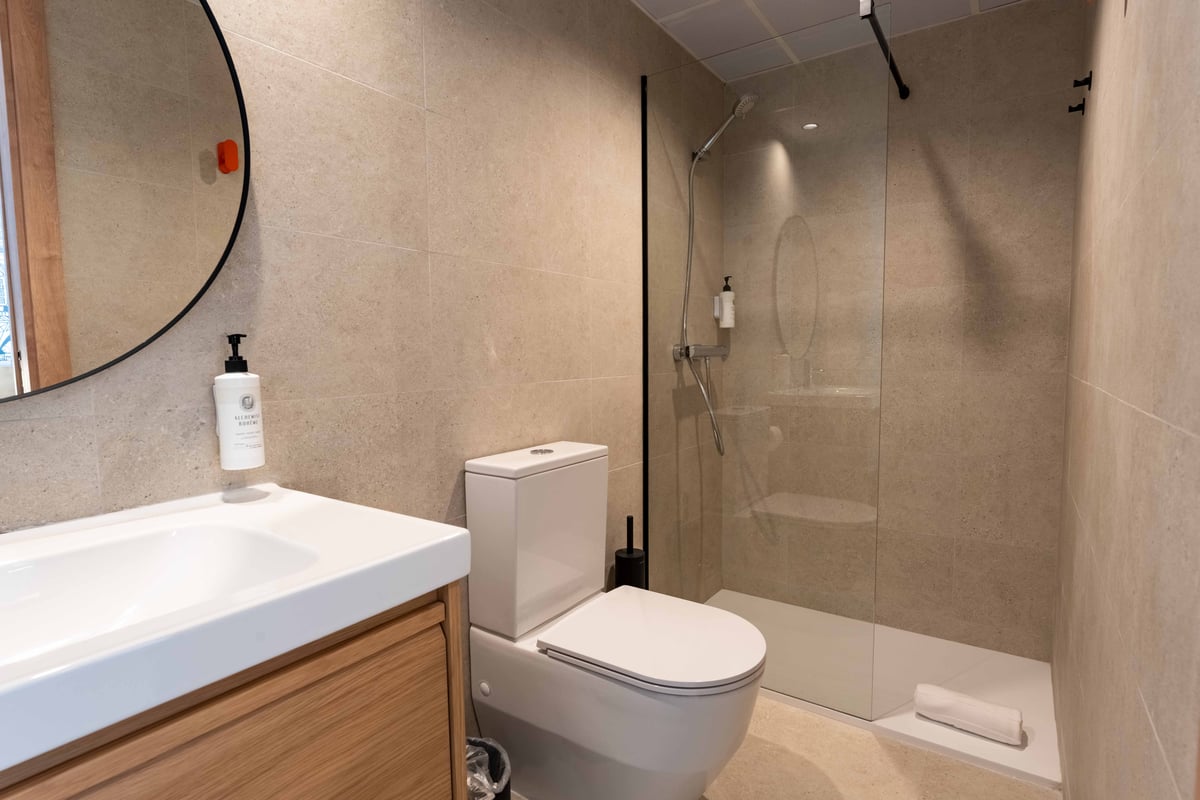Modern easyHotel Madrid Alcalá bathroom featuring a wooden vanity with a white sink, a black-framed round mirror, a white toilet, and a spacious walk-in shower with a glass partition and neutral-toned tiles.