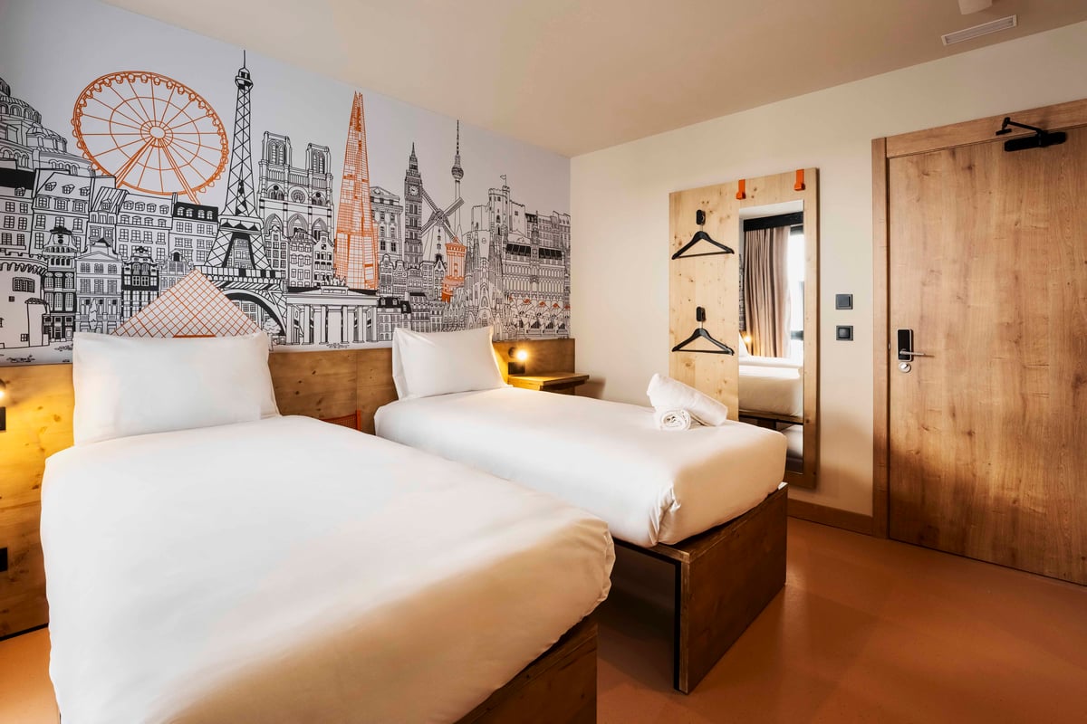 easyHotel Madrid Alcalá twin bedroom with two single beds, white bedding, and a prominent wall mural featuring a stylized cityscape. The minimalist room includes reading lamps and a mirror with hangers.