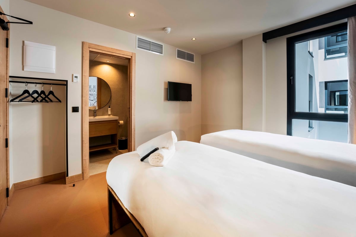 easyHotel Madrid Alcalá twin bedroom with two single beds, a wall-mounted TV, an open wardrobe with hangers, and an entrance to a modern private bathroom.