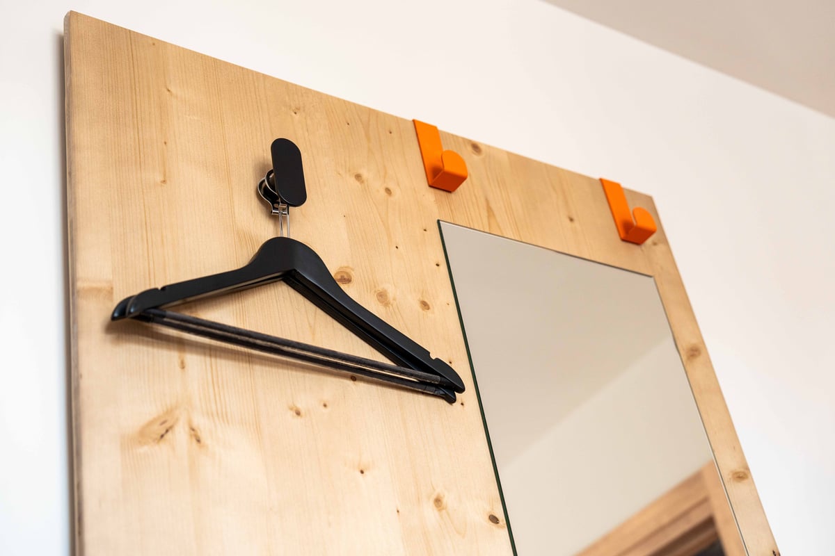Black clothing hanger on a minimalist wooden wall panel with orange hooks and a mirror in an easyHotel Madrid Alcalá room.
