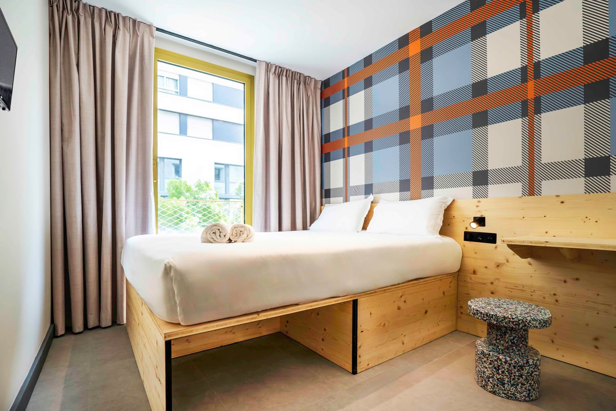 Simple double room with a wooden bed, plaid wallpaper, and a window overlooking a building.