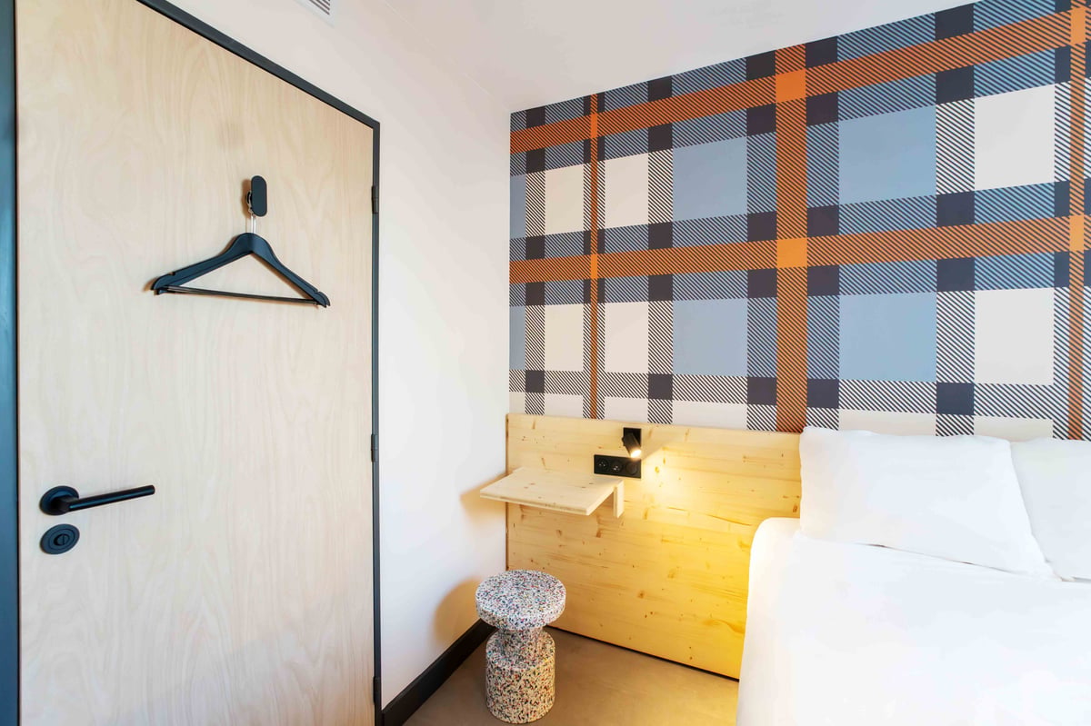 easyHotel Marseille Euromed accessible room featuring a vibrant plaid accent wall in blue, orange, and white, a light wooden headboard with an integrated shelf, white bed linen, and a wooden door with a clothes hanger.